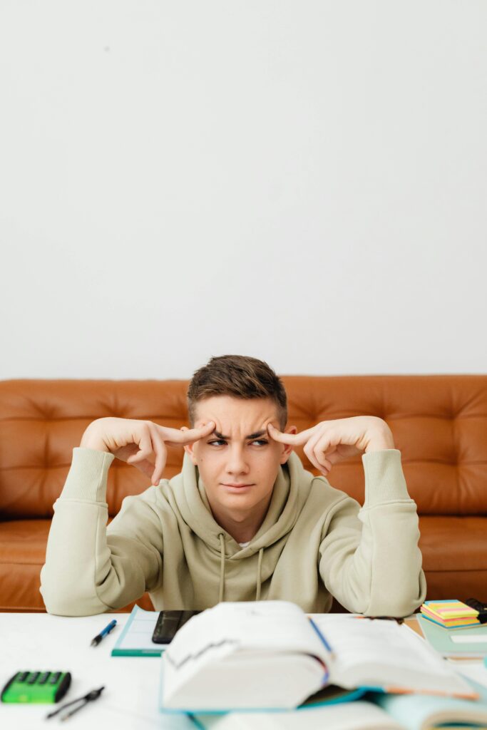 A teenager sits at home studying with a confused expression, surrounded by textbooks.